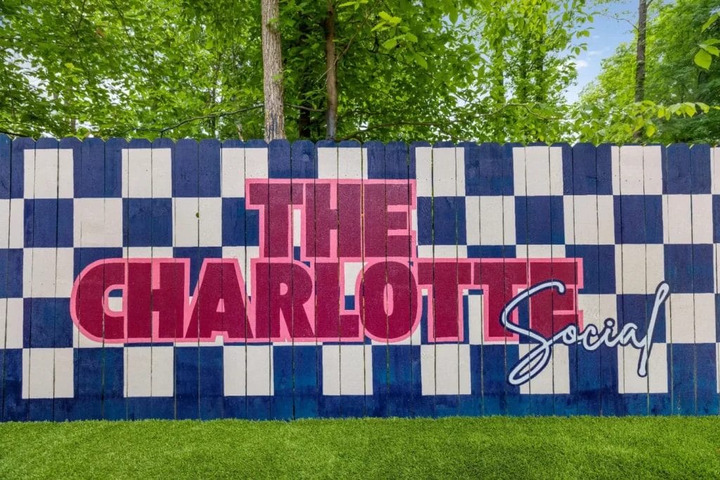 A painted fence features a blue and white checkerboard pattern with large red text reading "THE CHARLOTTE" and smaller cursive blue text below reading "Social," set in front of green trees and artificial grass.