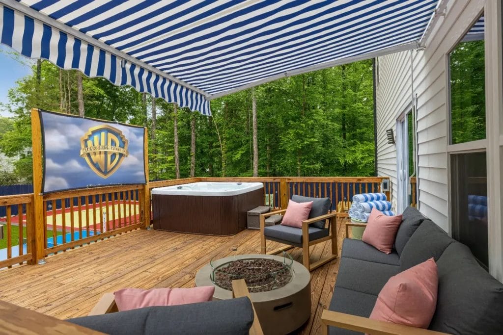 A cozy wooden deck with striped blue-and-white awning, gray outdoor sofas with pink pillows, a fire pit table, a hot tub, and a large Warner Bros. logo screen, overlooking a green wooded area.