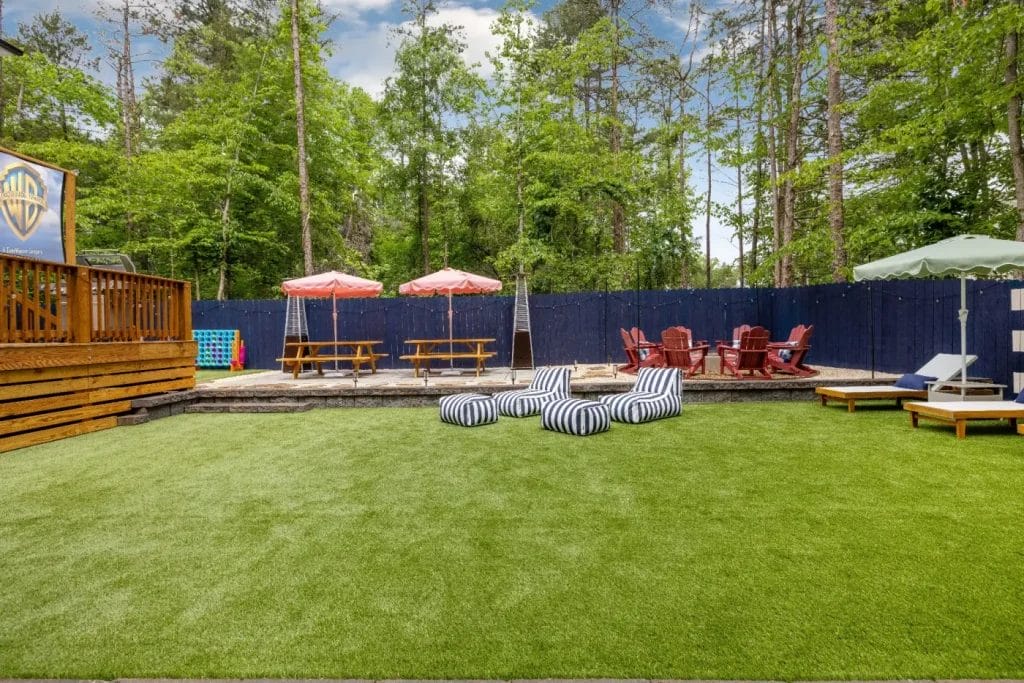 A spacious backyard features artificial grass, striped bean bag chairs, picnic tables with red umbrellas, Adirondack chairs around a fire pit, a wooden deck, and trees in the background.