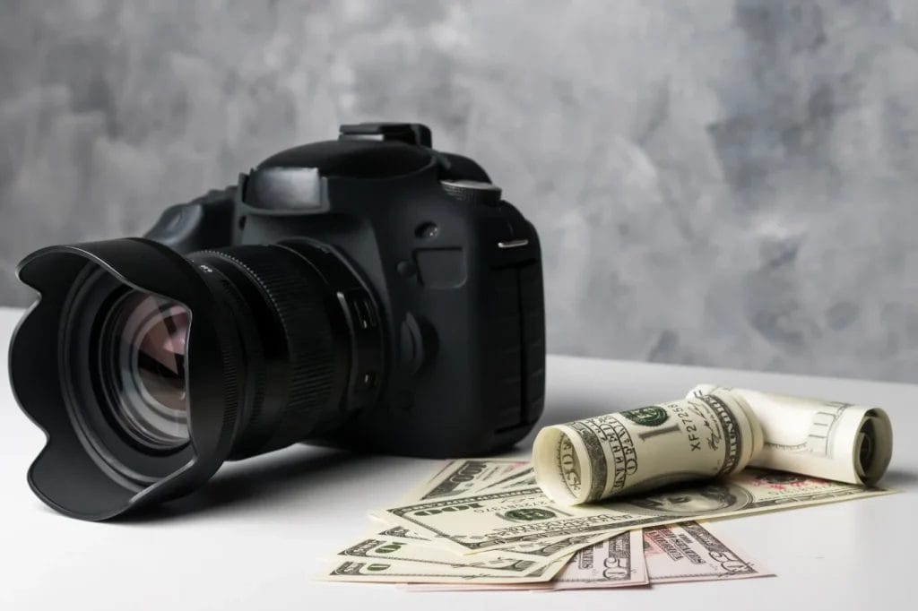 A black DSLR camera sits on a white surface next to a rolled-up stack of U.S. dollar bills, suggesting questions like "How Much Does Airbnb Photography Cost in Charlotte NC," with a gray textured background behind them.